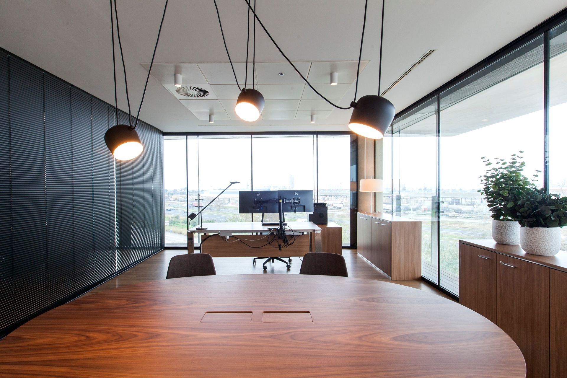 Modern office with large windows, wooden furniture, and hanging pendant lights.