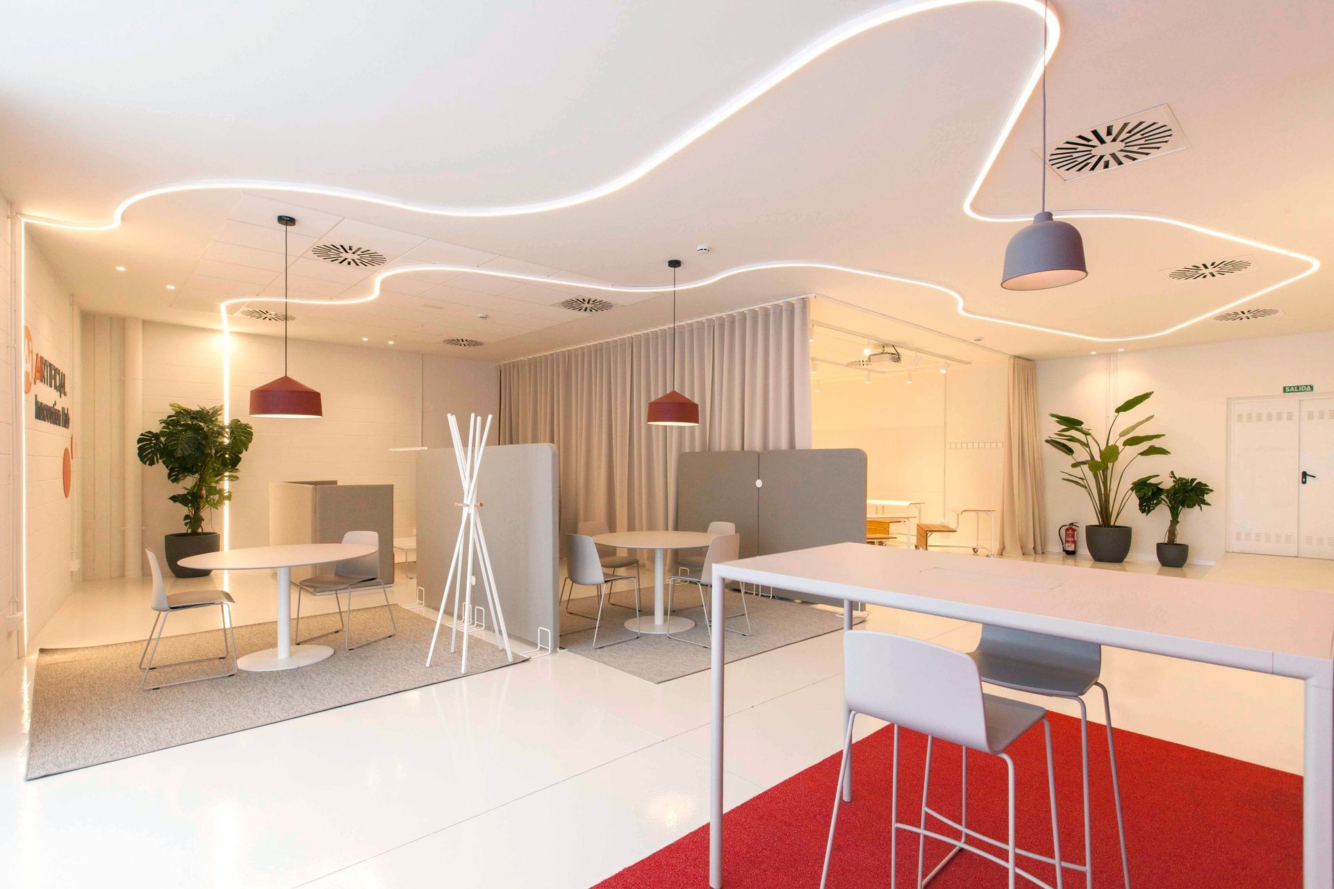 Modern office space with minimalist furniture, ceiling lights, plants, and partitioned seating areas.