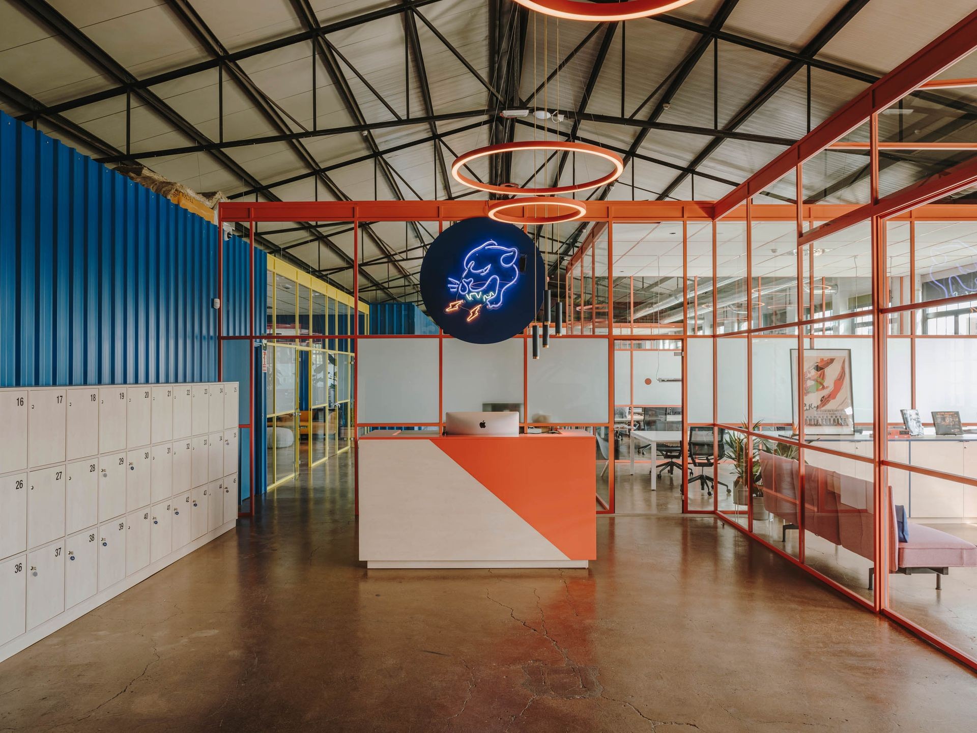 Modern office interior with glass walls, colorful lockers, and a reception desk under a neon sign.